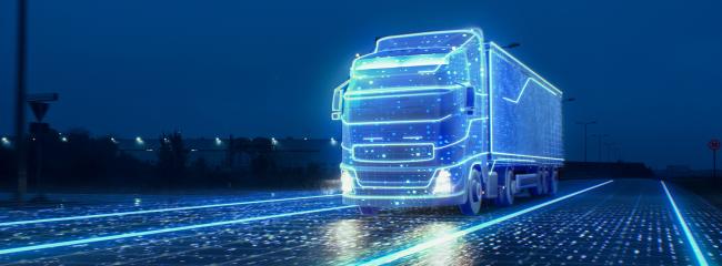 A futuristic, autonomous semitruck with a cargo trailer drives on a 4-lane highway at night. The truck and road each contain sensors that communicate in live time to allow the truck to drive safely. (Image courtesy of iStock.com / gorodenkoff)